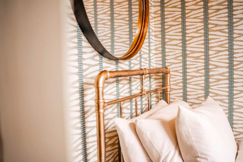 A close-up shot from a WildKind Interiors-styled bedroom in Anfield, Liverpool, showcasing a striking brass bedframe set against bespoke neutral and teal striped wallpaper. The image...