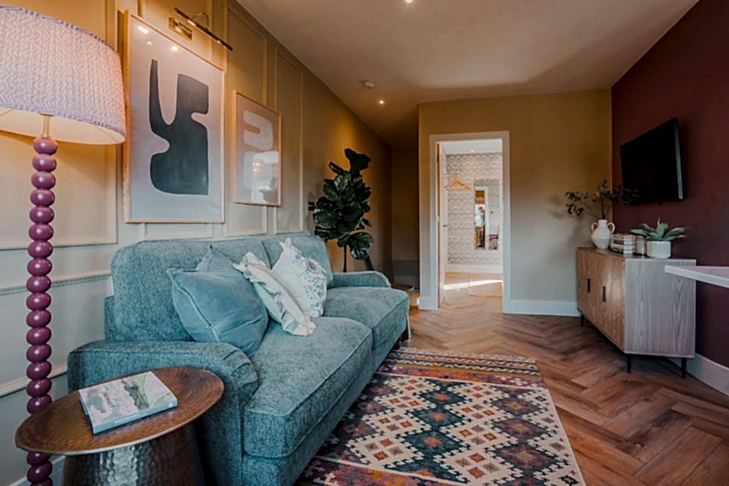 Explore this chic sitting room in Beckwithshaw, North Yorkshire serviced accommodation. Featuring a plush teal sofa with patterned and solid cushions against a paneled feature wall, a tall...