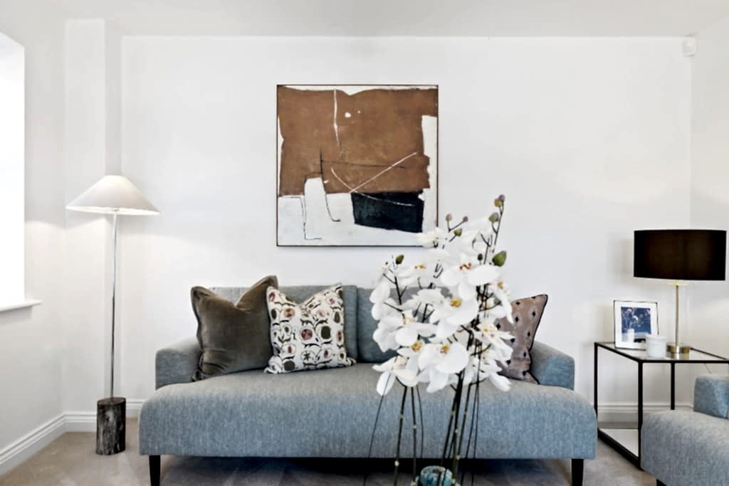 Living room with a soft blue sofa layered with patterned cushions, a floor lamp and a warm abstract artwork.