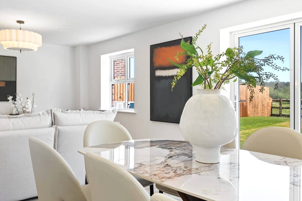 Open-plan dining area with a marble-effect table, cream chairs, a sculptural vase of greenery and a leafy garden view.