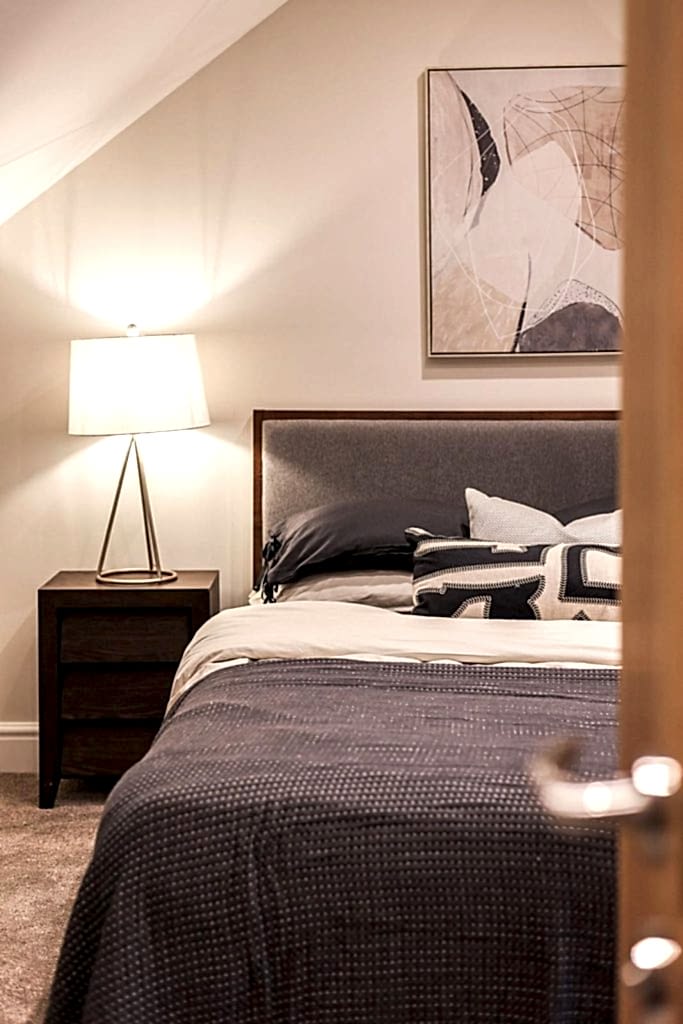 An inviting attic-style bedroom staged by WildKind Interiors in Huntingdon, York. This scene features a grey upholstered bed dressed in layered bedding, a dark wood bedside table...