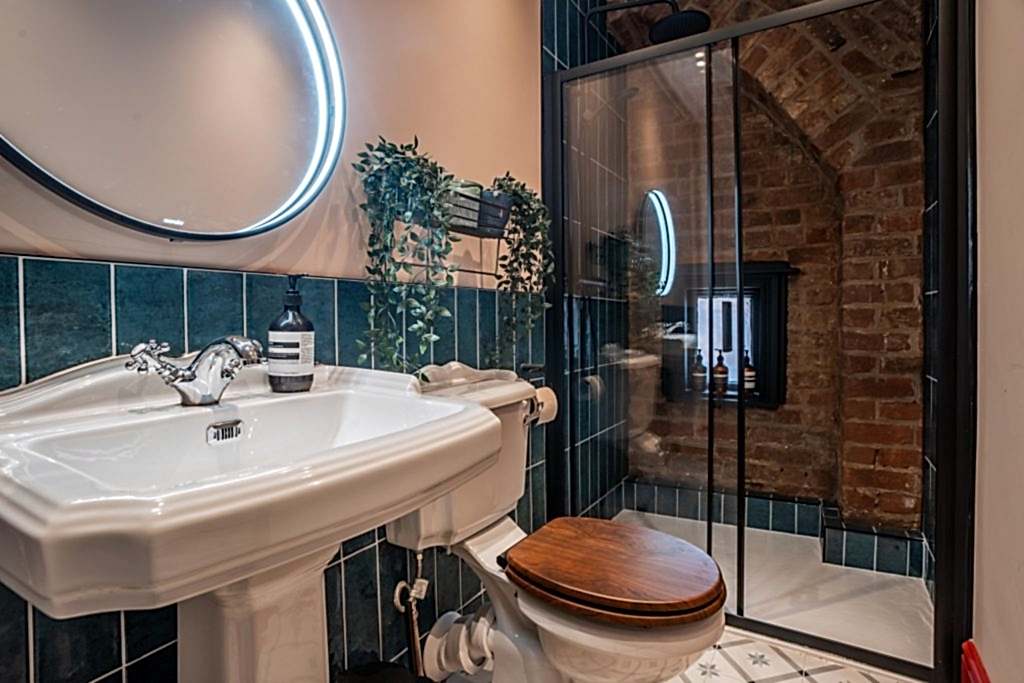 A characterful bathroom in Magazine Lane serviced accommodation, The Wirral, styled by WildKind Interiors. This design features a traditional white pedestal sink beneath an illuminated...