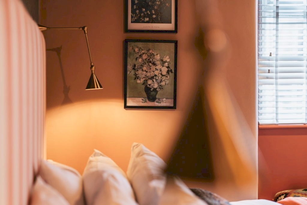 Bedroom detail with a striped headboard, a brass wall light and a framed floral print in warm evening light.