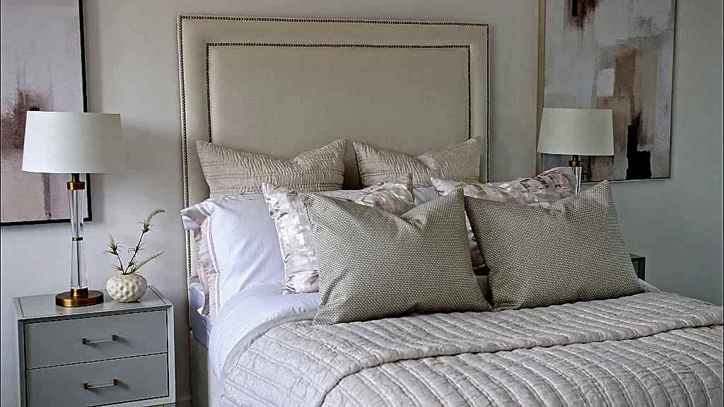 Bedroom styled with an upholstered headboard, layered cream cushions and matching bedside lamps beneath soft abstract artwork.