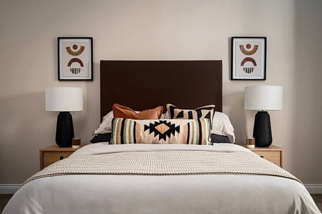 A modern bedroom expertly staged by WildKind Interiors, featuring a dark upholstered headboard, layered decorative pillows with geometric patterns, matching bedside lamps on wooden...