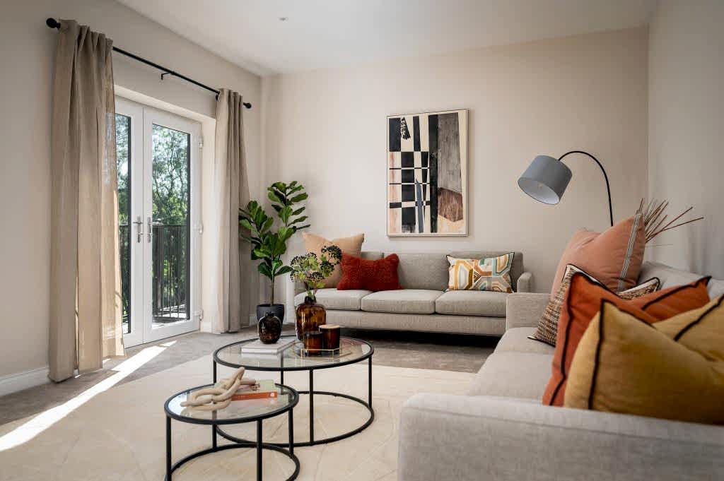 A bright, airy living room staged by WildKind Interiors featuring neutral-toned sofas layered with colorful accent cushions, nested glass coffee tables with decorative vases, a floor lamp...