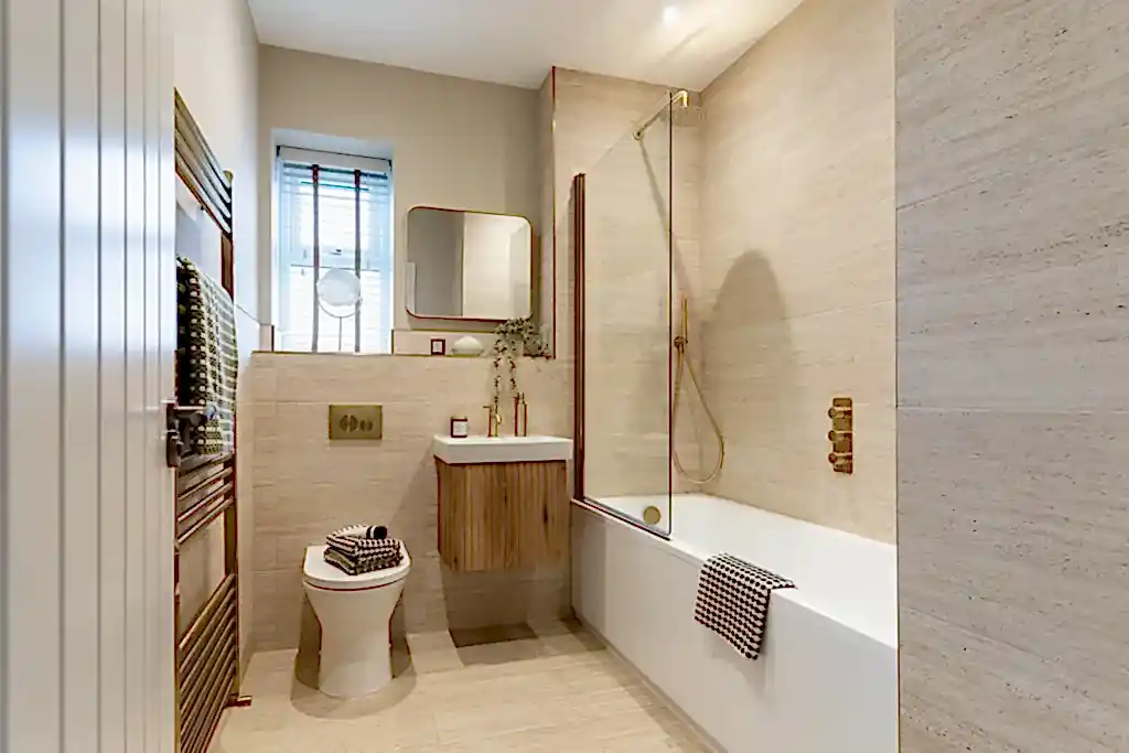 Bathroom with travertine effect tiles, a walk in shower over the bath and brass fittings.